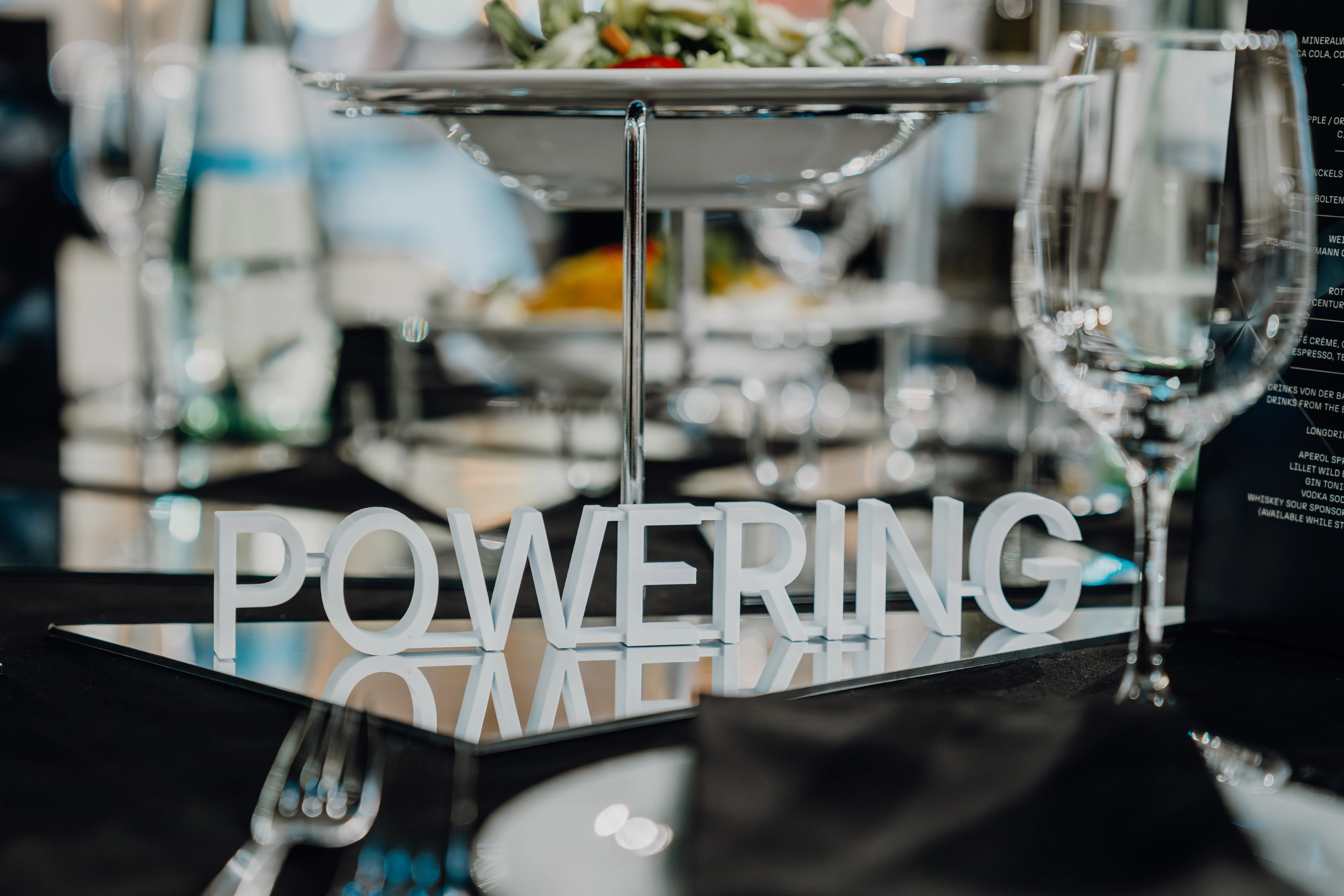 Weiße 3D-gedruckte Buchstaben bilden das Wort POWERING auf einer gedeckten Eventtafel.